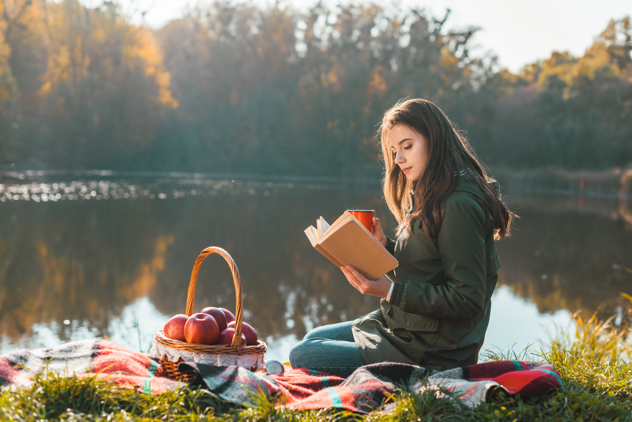 beautiful young woman with coffee cup reading book on blanket near pond in park